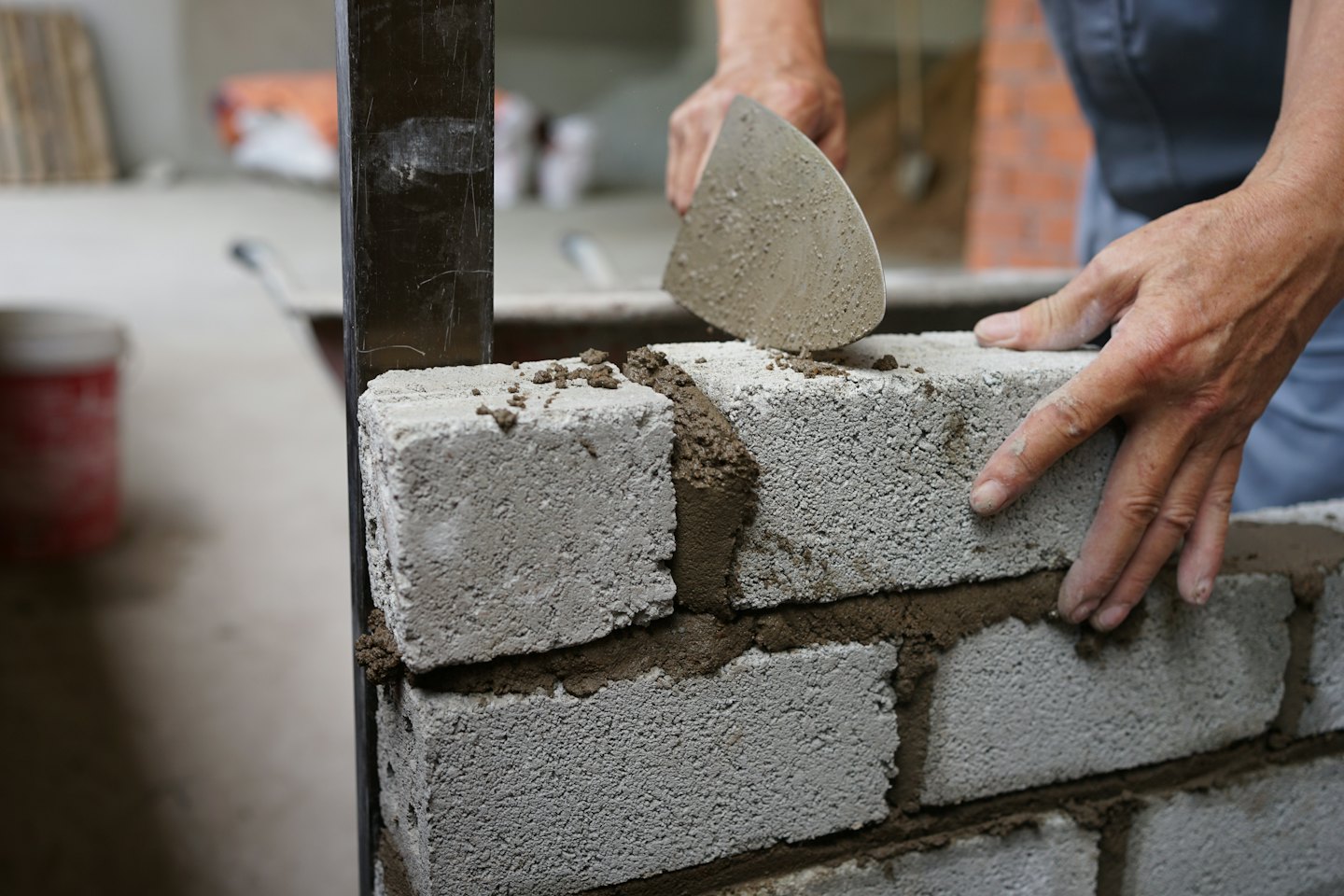 Handen van een bouwvakker die mortel en betonblokken legt voor een muur.